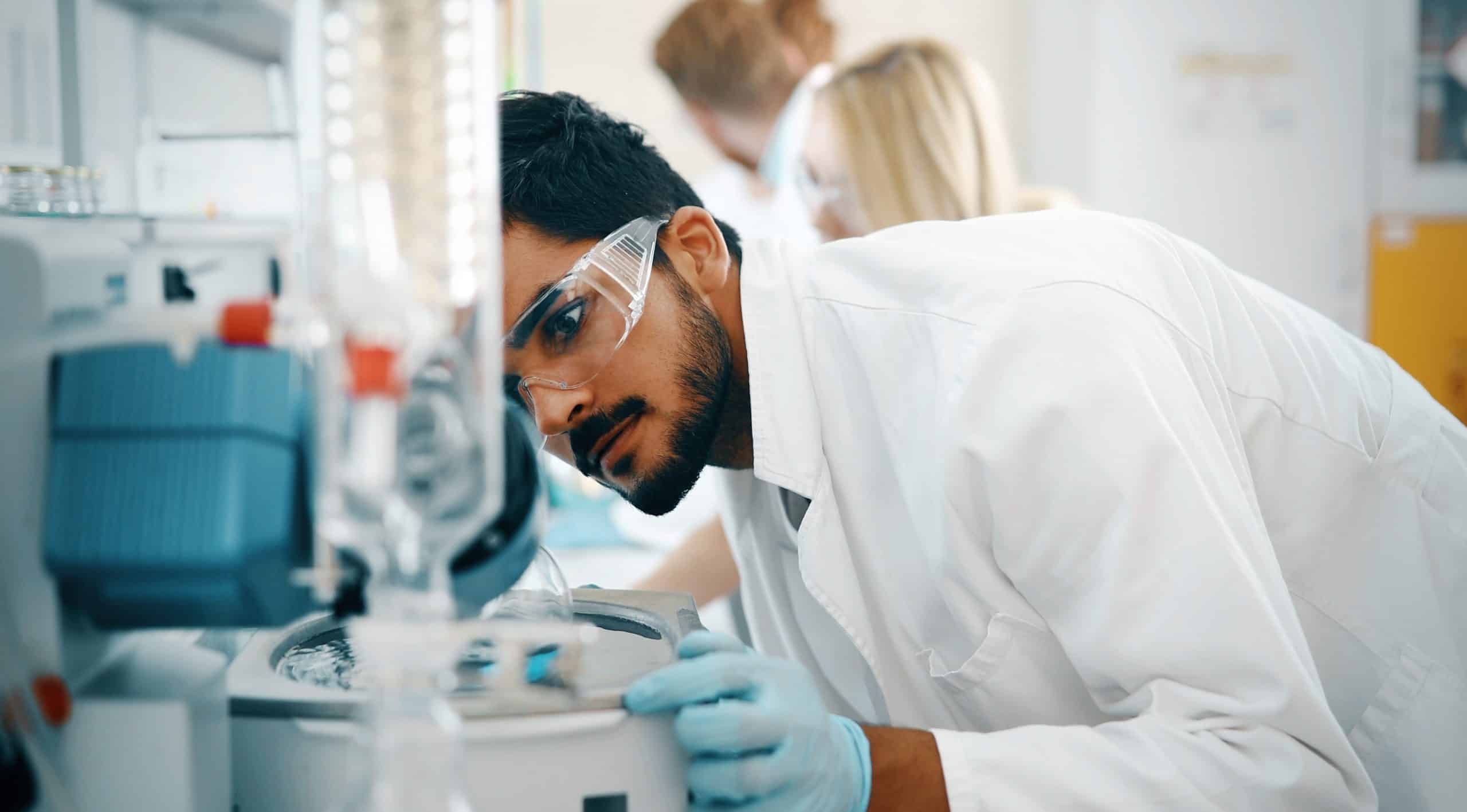 Scientist wearing goggles examines equipment in a lab.