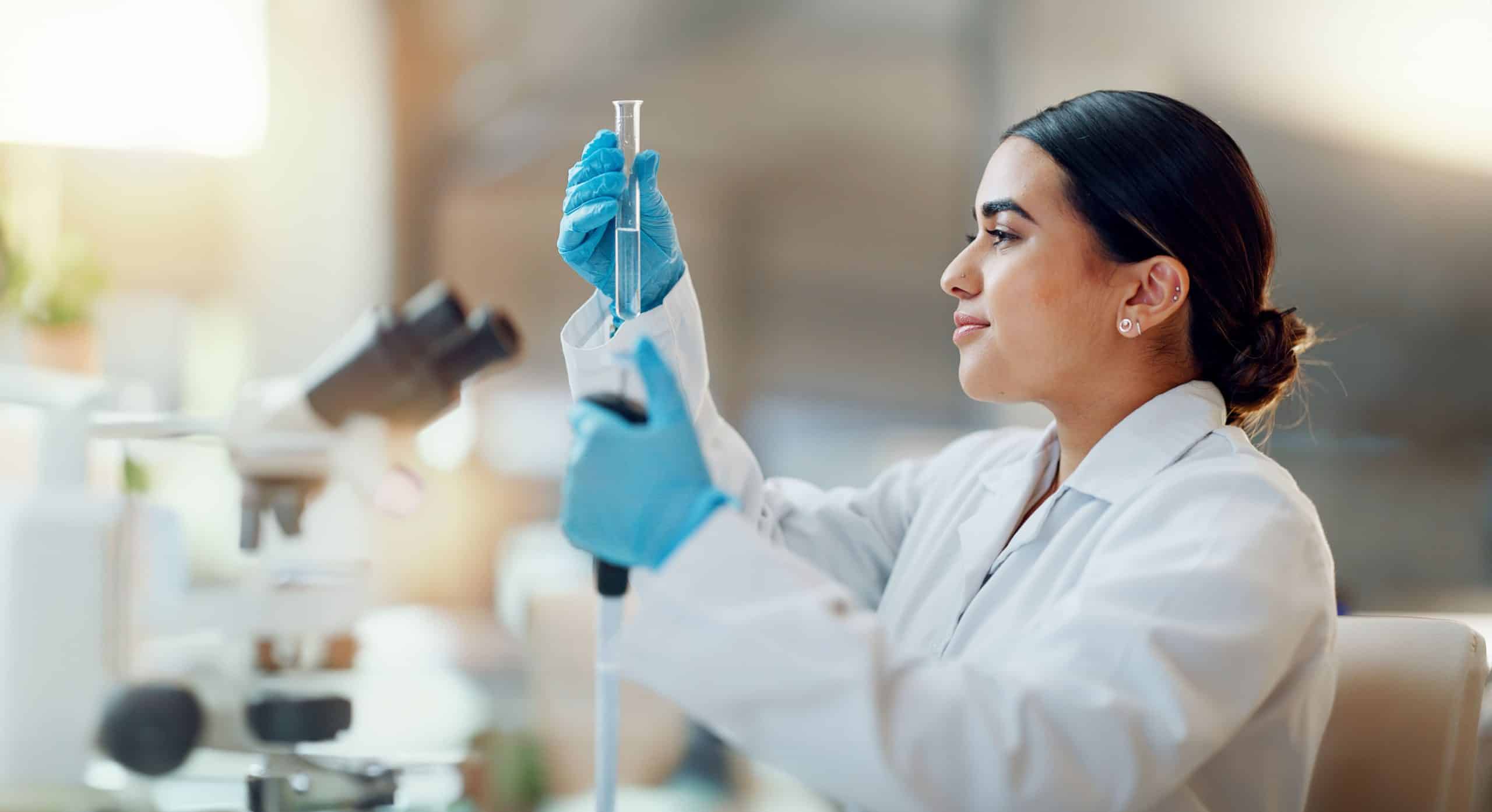 ChemicalCompanies Scientist in a lab holding a test tube and pipette, with a microscope in the foreground.